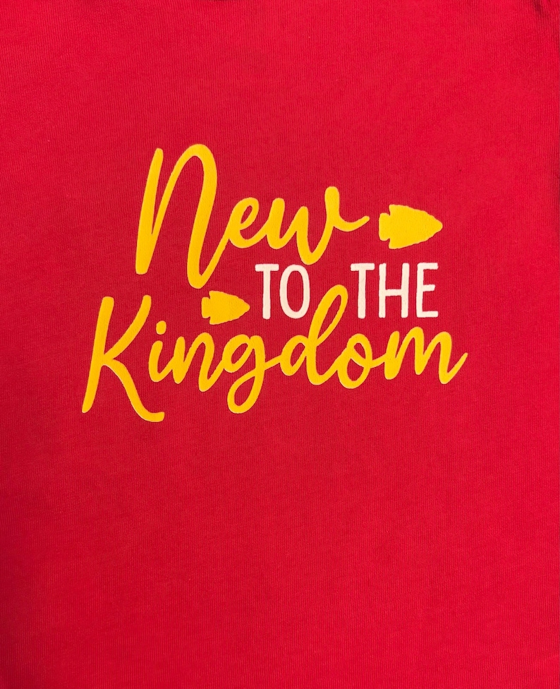 May include: A red t-shirt with the words "New to the Kingdom" in yellow script and white block letters. The design includes yellow arrowhead graphics. The shirt is a solid color.