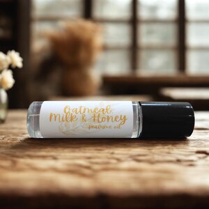 Puede incluir: Un frasco roll-on de vidrio transparente con tapa negra, etiquetado "Oatmeal Milk & Honey perfume oil" en escritura dorada. El frasco descansa sobre una superficie de madera, con un fondo borroso con flores y una ventana.