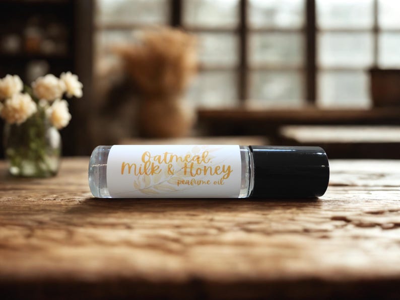 Puede incluir: Un frasco roll-on de vidrio transparente con tapa negra, etiquetado "Oatmeal Milk & Honey perfume oil" en escritura dorada. El frasco descansa sobre una superficie de madera, con un fondo borroso con flores y una ventana.