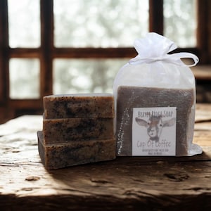 May include: A stack of three rectangular soaps with dark specks, next to a clear bag of soap. The bag has a label that reads "BRAYER RIDGE SOAP" with a goat illustration and "Cup Of Coffee". The soaps are on a rustic wooden surface.