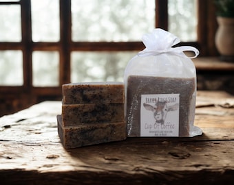 May include: A stack of three rectangular soaps with dark specks, next to a clear bag of soap. The bag has a label that reads "BRAYER RIDGE SOAP" with a goat illustration and "Cup Of Coffee". The soaps are on a rustic wooden surface.
