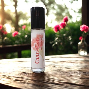 May include: A clear glass roll-on bottle with a black cap, labeled "Carnation perfume oil" in a script font with a floral design. The bottle sits on a wooden surface, with blurred pink flowers and greenery in the background.