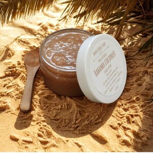 May include: A jar of brown sugar scrub with a white lid, labeled "Bahama Coconut" and a small wooden spoon. The jar is open, revealing the scrub's texture. The items are on a sandy surface, with palm fronds in the background.