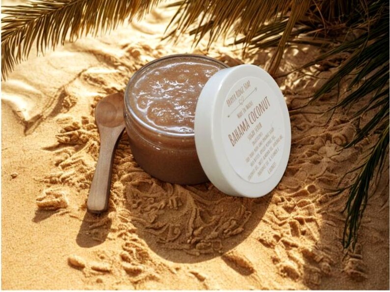 May include: A jar of brown sugar scrub with a white lid, labeled "Bahama Coconut" and a small wooden spoon. The jar is open, revealing the scrub's texture. The items are on a sandy surface, with palm fronds in the background.