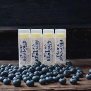 May include: Four cylindrical lip balm tubes with white caps and labels featuring the text Maine Blueberry Lip Balm in blue script, arranged in a row behind a scattered pile of fresh, dark blue blueberries on a dark wooden surface.