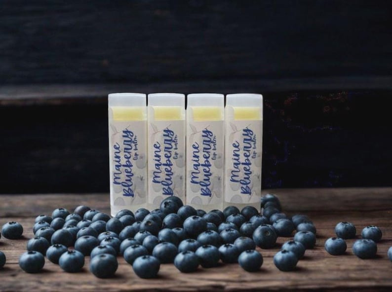 May include: Four cylindrical lip balm tubes with white caps and labels featuring the text Maine Blueberry Lip Balm in blue script, arranged in a row behind a scattered pile of fresh, dark blue blueberries on a dark wooden surface.