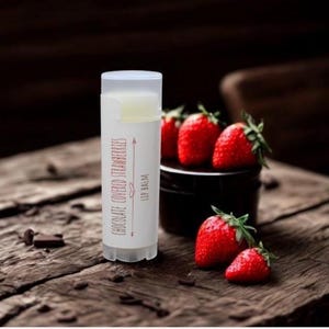May include: A white lip balm tube with the text "Chocolate Covered Strawberries" and "Lip Balm" in red. Fresh red strawberries are next to the tube on a wooden surface. Chocolate pieces are scattered nearby.