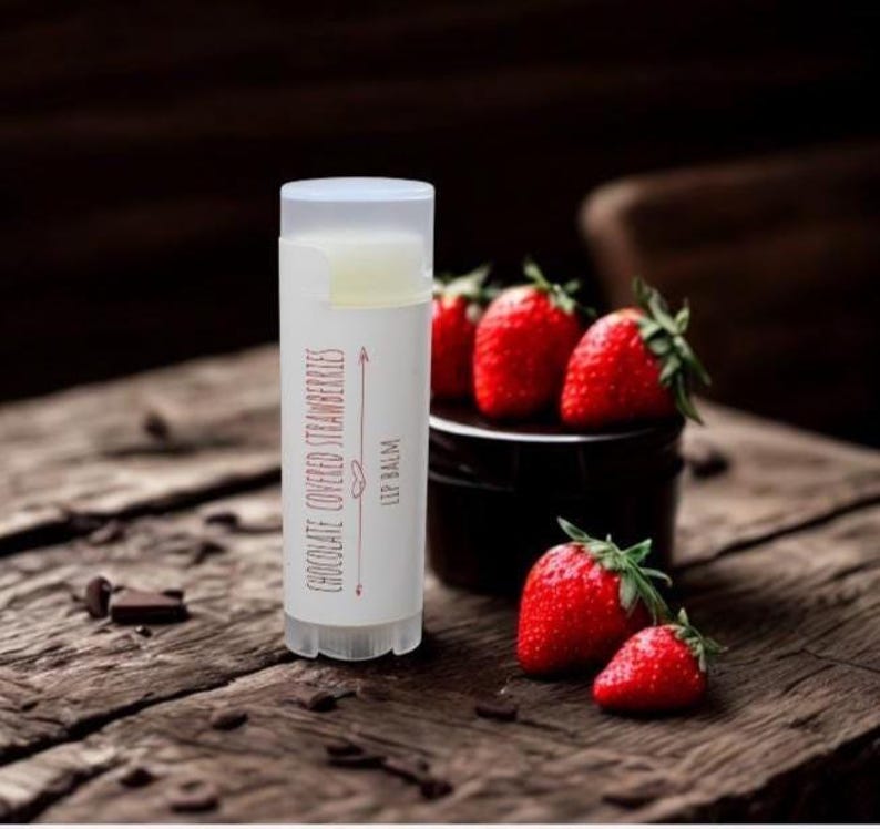 May include: A white lip balm tube with the text "Chocolate Covered Strawberries" and "Lip Balm" in red. Fresh red strawberries are next to the tube on a wooden surface. Chocolate pieces are scattered nearby.