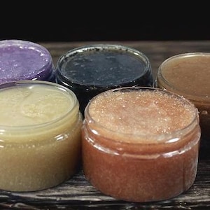 May include: Five open jars of body scrub with various colors and textures. The scrubs range in color from purple to brown, with visible granules. The jars are clear, and the scrubs are sitting on a wooden surface.