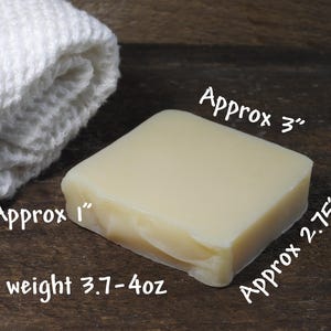 May include: A square bar of light-colored soap, approximately 3 inches by 2.75 inches, with a net weight of 3.7-4 oz, next to a rolled white washcloth. The soap has a smooth surface and a slightly translucent appearance.