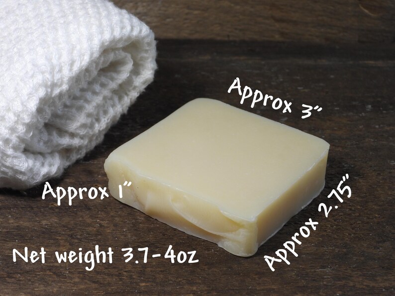 May include: A square bar of light-colored soap, approximately 3 inches by 2.75 inches, with a net weight of 3.7-4 oz, next to a rolled white washcloth. The soap has a smooth surface and a slightly translucent appearance.