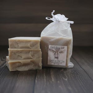 May include: A stack of three light-colored soap bars next to a bag of soap. The bag is clear with a white ribbon and a label that reads "Beaver Ridge Soap Cambrian Blue Clay". The soap bars have a textured appearance.