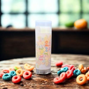 May include: A clear lip balm tube with the text "fruit loops" in colorful letters. The tube is surrounded by colorful, circular cereal pieces on a wooden surface. The background is blurred, showing a window and some fruit.