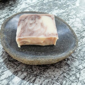 May include: A square bar of soap with a cream and mauve marbled pattern resting on a dark gray, oval-shaped stone dish. The soap has a smooth, solid texture and sits centered within the shallow, natural-looking basin of the stone.