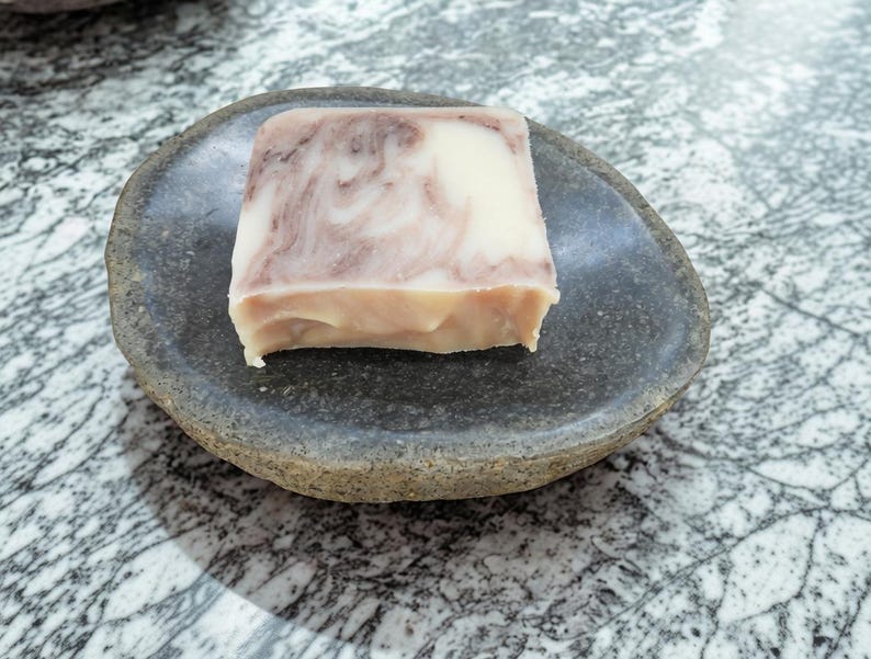 May include: A square bar of soap with a cream and mauve marbled pattern resting on a dark gray, oval-shaped stone dish. The soap has a smooth, solid texture and sits centered within the shallow, natural-looking basin of the stone.