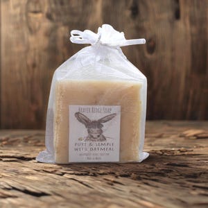 May include: A square bar of beige soap in a white mesh bag tied with a ribbon. The soap has a label with a rabbit illustration and the text "PURE & SIMPLE WITH OATMEAL". The background is a rustic wooden surface.