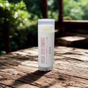 May include: A white lip balm tube with a clear cap, labeled "Chocolate Covered Strawberries Lip Balm." The tube is on a weathered wooden surface, with a blurred green background.
