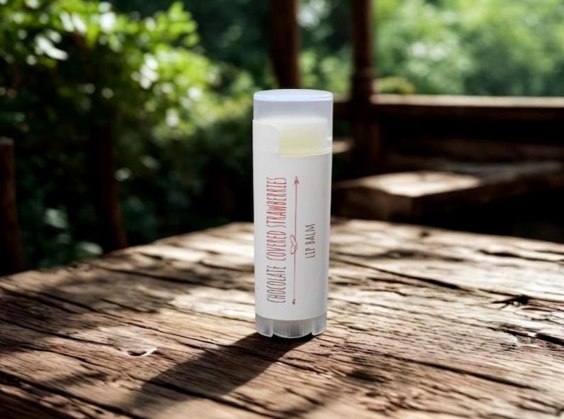 May include: A white lip balm tube with a clear cap, labeled "Chocolate Covered Strawberries Lip Balm." The tube is on a weathered wooden surface, with a blurred green background.