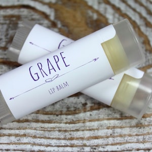 May include: Two clear lip balm tubes with white labels. The label on the top tube says "GRAPE LIP BALM" in purple text with a small heart and arrows on either side.
