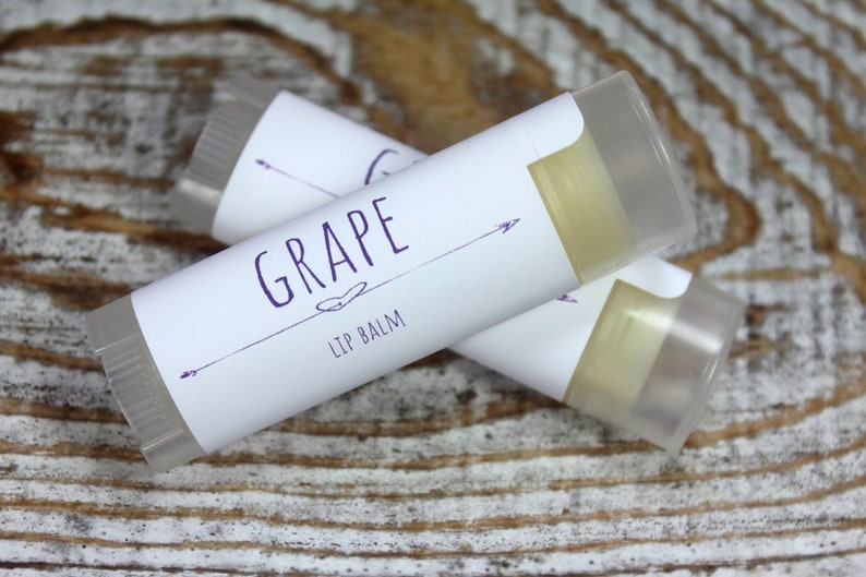 May include: Two clear lip balm tubes with white labels. The label on the top tube says "GRAPE LIP BALM" in purple text with a small heart and arrows on either side.