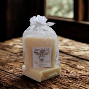 May include: Two bars of soap, one stacked on top of the other, are presented in a sheer white bag tied with a silver ribbon. The soap bars are light beige with a label that reads "BRAYER RIDGE SOAP" and "Bourbon Tobacco". The background is a rustic wooden surface.