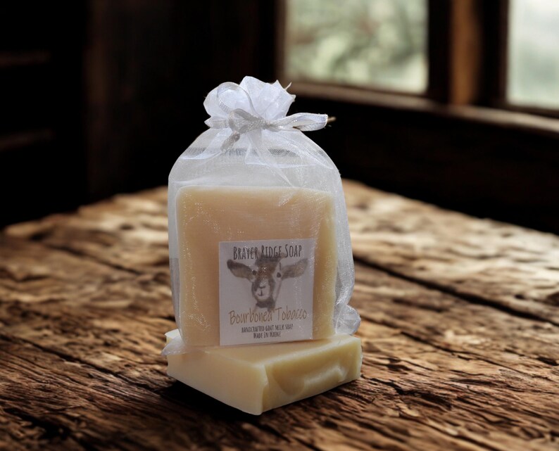 May include: Two bars of soap, one stacked on top of the other, are presented in a sheer white bag tied with a silver ribbon. The soap bars are light beige with a label that reads "BRAYER RIDGE SOAP" and "Bourbon Tobacco". The background is a rustic wooden surface.