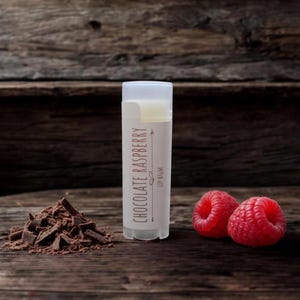 May include: A lip balm tube with the words "Chocolate Raspberry" printed on it, next to a pile of chocolate shavings and two fresh raspberries. The lip balm is in a clear tube with a white cap.