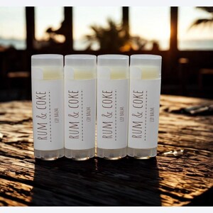 May include: Four lip balm tubes with white labels and the text "RUM & COKE" in brown. The lip balms are arranged on a wooden surface, with a blurred background of a beach scene. The lip balms are a cosmetic product.