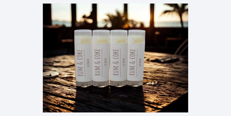 May include: Four lip balm tubes with white labels and the text "RUM & COKE" in brown. The lip balms are arranged on a wooden surface, with a blurred background of a beach scene. The lip balms are a cosmetic product.