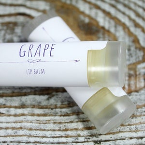 May include: Two white tubes of lip balm with a white label that says "GRAPE LIP BALM" in purple text. The tubes are on a light brown wooden surface.