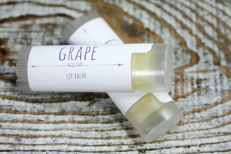 May include: Two white tubes of lip balm with a white label that says "GRAPE LIP BALM" in purple text. The tubes are on a light brown wooden surface.