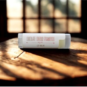 May include: A clear lip balm tube with a white label that reads "Chocolate Covered Strawberries" in a brown script. The lip balm is on a wooden surface, with sunlight streaming in from a window in the background.