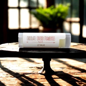 May include: A lip balm tube with a white label that reads "Chocolate Covered Strawberries" in a decorative font. The lip balm is on a dark wooden table with a blurred background of a window and greenery.