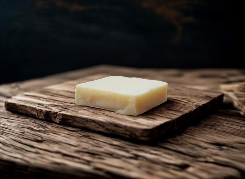 May include: A square bar of light-colored soap rests on a weathered wooden surface. The soap has a slightly irregular shape, suggesting a handmade quality. The wood grain is prominent, adding a rustic feel.