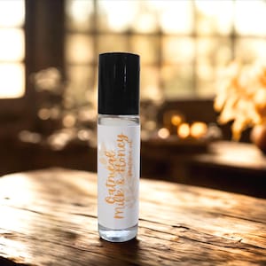Puede incluir: Un frasco de aceite de perfume de vidrio transparente con una tapa negra. La etiqueta dice "Oatmeal Milk & Honey perfume oil" en una fuente cursiva. El fondo es una superficie de madera borrosa y una vista de enfoque suave de una habitaci&oacute;n.