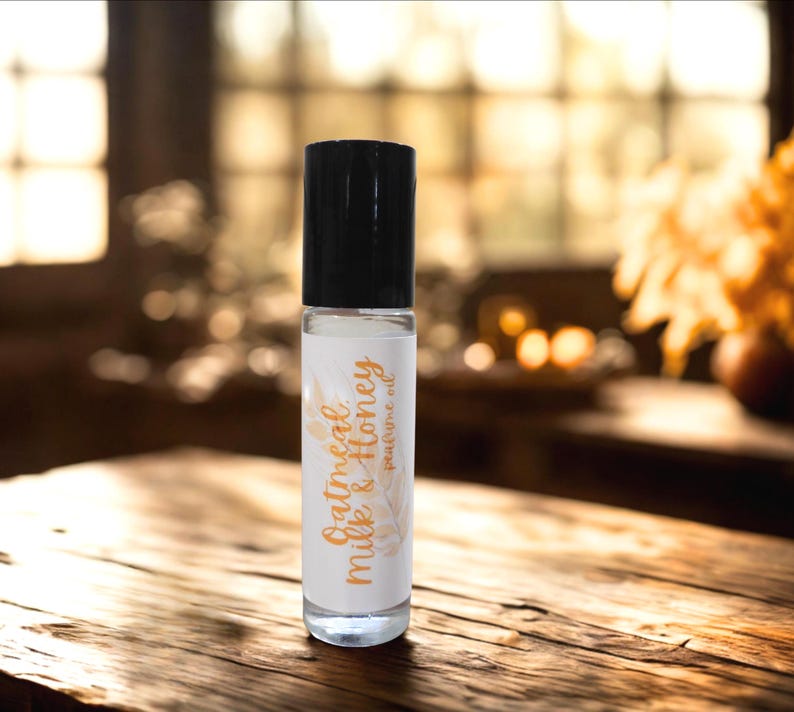Puede incluir: Un frasco de aceite de perfume de vidrio transparente con una tapa negra. La etiqueta dice "Oatmeal Milk & Honey perfume oil" en una fuente cursiva. El fondo es una superficie de madera borrosa y una vista de enfoque suave de una habitaci&oacute;n.