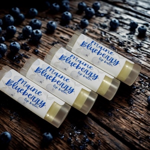 May include: Four cylindrical lip balm tubes with white labels featuring blue text that reads Maine Blueberry lip balm. The tubes are arranged diagonally, each containing a pale yellow balm, and are surrounded by scattered fresh blueberries.