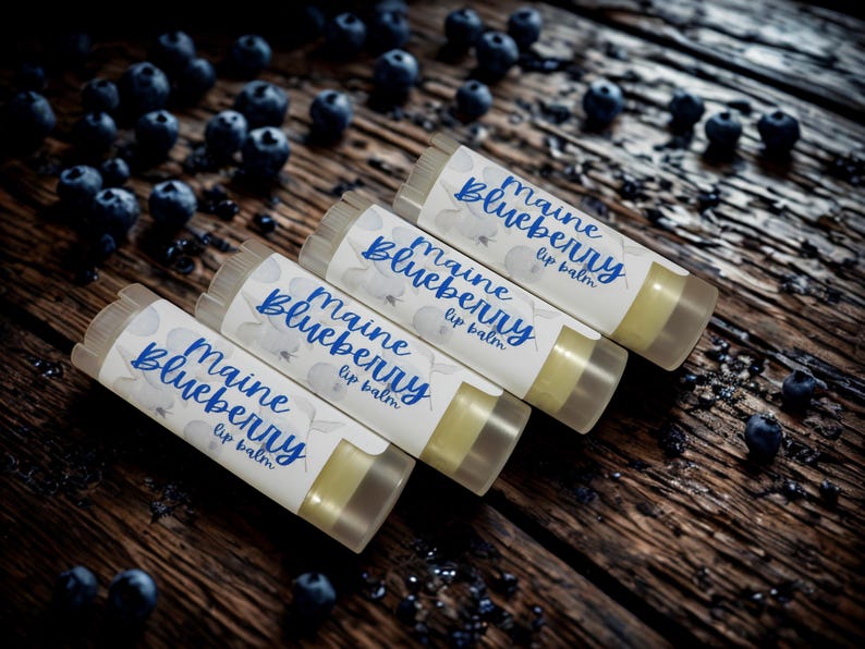 May include: Four cylindrical lip balm tubes with white labels featuring blue text that reads Maine Blueberry lip balm. The tubes are arranged diagonally, each containing a pale yellow balm, and are surrounded by scattered fresh blueberries.