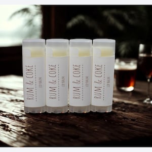 May include: Four lip balm tubes with the text "RUM & COKE" on a white label. The lip balms are on a dark wooden surface. The background includes a window and glasses of a dark beverage.