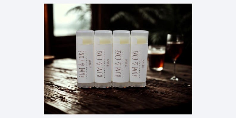 May include: Four lip balm tubes with the text "RUM & COKE" on a white label. The lip balms are on a dark wooden surface. The background includes a window and glasses of a dark beverage.