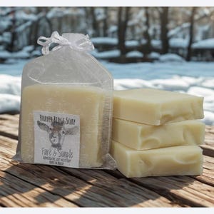 Puede incluir: Una bolsa transparente contiene una barra grande de jab&oacute;n con una etiqueta que dice "Beaver Ridge Soap" y "Fine & Simple". Al lado, tres barras rectangulares de jab&oacute;n est&aacute;n apiladas sobre una superficie de madera, con un fondo nevado.