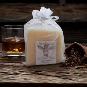 May include: A bar of BRAYER RIDGE SOAP, Bourboned Tobacco, in a clear cellophane bag tied with a white ribbon. A glass of amber liquid with ice and a small leather pouch with tobacco leaves are also in the image.
