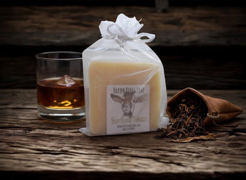 May include: A bar of BRAYER RIDGE SOAP, Bourboned Tobacco, in a clear cellophane bag tied with a white ribbon. A glass of amber liquid with ice and a small leather pouch with tobacco leaves are also in the image.
