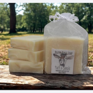 Puede incluir: Una pila de tres jabones rectangulares de color claro junto a una barra de jab&oacute;n m&aacute;s grande en una bolsa de malla. La bolsa tiene una etiqueta que dice "Beaver Ridge Soap" con una ilustraci&oacute;n de cabra y las palabras "Pure & Simple".