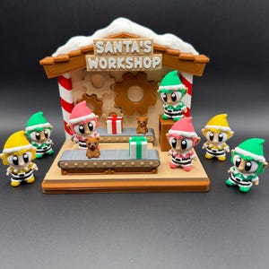 May include: A festive Santa's Workshop scene featuring a sign with the text "SANTA'S WORKSHOP". The scene includes a conveyor belt with wrapped gifts and toy bears, surrounded by elf figurines wearing green, pink, and yellow hats.