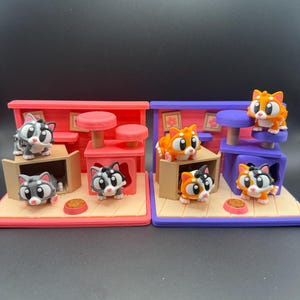 May include: Two miniature cat playhouses, one red and one purple, each with several cartoon cat figurines. The cats are in various poses, some in boxes, others on platforms. Each playhouse has a food bowl.
