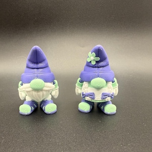 Puede incluir: Dos figuras de gnomos caprichosos. Cada gnomo tiene un sombrero morado, una barba blanca y ropa verde y blanca. Un gnomo tiene una flor en su sombrero. Los gnomos miden aproximadamente 7,6 cm de altura.