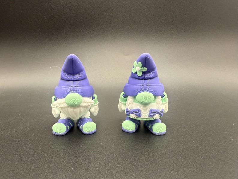Puede incluir: Dos figuras de gnomos caprichosos. Cada gnomo tiene un sombrero morado, una barba blanca y ropa verde y blanca. Un gnomo tiene una flor en su sombrero. Los gnomos miden aproximadamente 7,6 cm de altura.