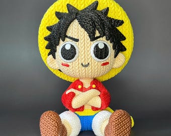 May include: A knitted Luffy figurine, featuring a yellow straw hat and black spiky hair. The figure wears a red jacket, blue shorts, and brown boots. The character has large, expressive eyes and is in a seated position.