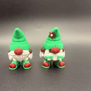 Puede incluir: Dos figuras de gnomos caprichosos. Cada gnomo tiene un gorro puntiagudo verde, una nariz roja y una barba blanca. Llevan zapatos y mitones rojos. Un gnomo tiene una flor en su sombrero. Para decoraci&oacute;n navide&ntilde;a o de jard&iacute;n.